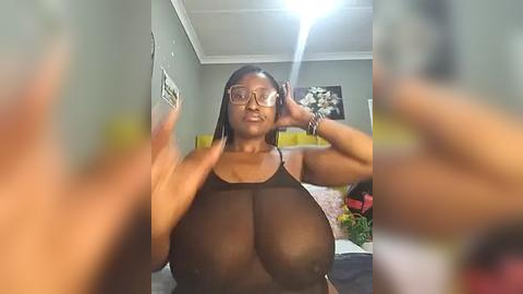 Media: Video of a Black woman with a large breast size, wearing a sheer black top, taking a selfie in a dimly lit room.