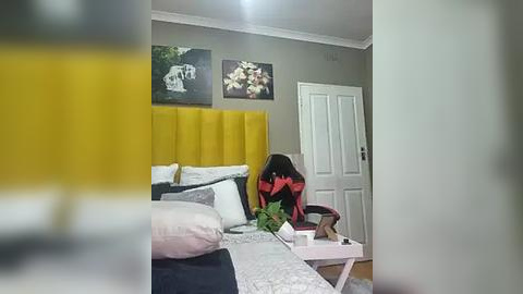 Media: Video of a modern bedroom with a yellow upholstered headboard, white door, gray walls, floral artwork, and a person sitting on a bed with a red and black jacket.