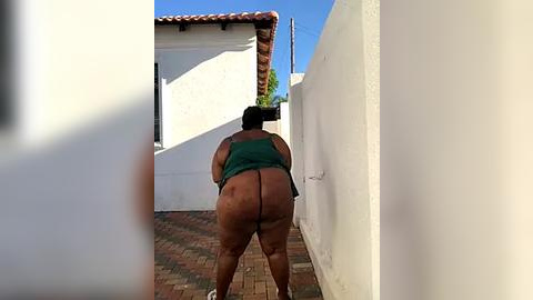 Media: Video of a dark-skinned woman with a large physique, wearing only a green top, standing barefoot on a brick patio, leaning against a white wall.