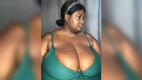 Media: A video of a plus-sized Black woman with a dark complexion, wearing a green camisole with thin straps, and a patterned headband. She stands against a neutral, blurred background, emphasizing her large breasts.