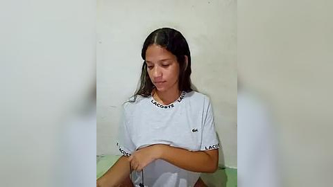 Media: Video of a young Hispanic woman with long black hair, wearing a white Lacoste t-shirt, sitting on a light green surface against a plain beige wall. She looks down, appearing contemplative.