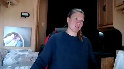 Media: Video of a woman with light skin, light brown hair, and blue sweater, standing in a dimly lit RV kitchen with beige cabinets, a flat-screen TV displaying a flower image, and a cluttered countertop.