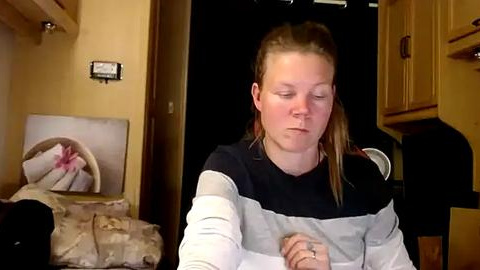 Media: A video of a fair-skinned woman with blonde hair tied back, wearing a striped sweater, sitting in a dimly lit kitchen. The background includes a closed door, wooden cabinets, and a framed photo on the wall.