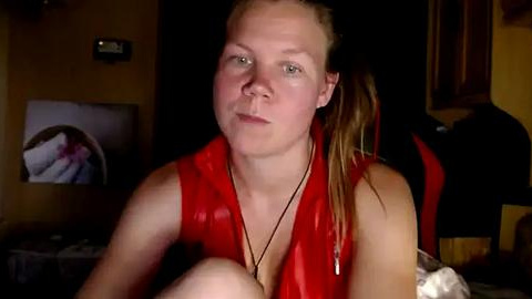 Media: A video of a young woman with fair skin, light brown hair tied back, wearing a red sleeveless top, seated indoors with dim lighting, looking relaxed.