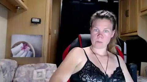 Media: Video of a fair-skinned woman with long brown hair in a ponytail, wearing a black lace camisole, seated in a small RV. Background includes a kitchen with wooden cabinets, a closed door, and a framed picture.