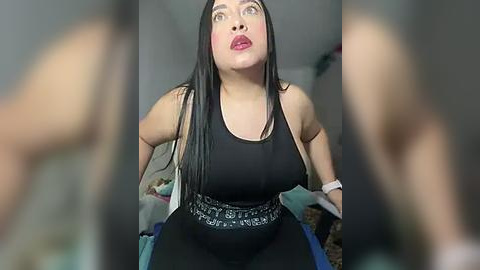 Media: Video of a plus-sized Latina woman with long black hair, wearing a black tank top and black skirt, sitting on a bed, looking surprised, with blurred figures in the background.