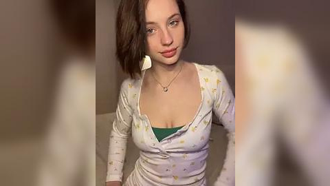 Media: Video of a young Caucasian woman with medium-length brown hair, fair skin, and green eyes, wearing a white button-up shirt with yellow polka dots over a green bra, seated in a dimly lit room.