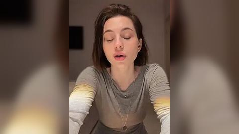 Media: Video of a young woman with light skin, closed eyes, and a slightly open mouth, wearing a grey and yellow striped shirt, leaning forward, blurred background.