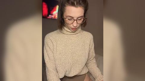 Media: Video of a young woman with fair skin, light brown hair in a ponytail, wearing glasses and a beige turtleneck sweater. She is in a dimly lit room with a TV displaying a red, glowing screen in the background.