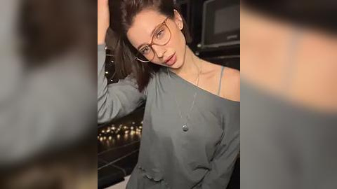 Media: A video of a fair-skinned woman with glasses and a grey off-shoulder top, standing in a dimly-lit room with a blurred background of shelves and a microwave.