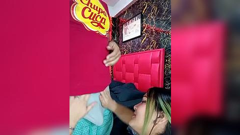 Media: Video of a young woman with green hair, wearing a green sweater, lying on a red bed with a red quilt, surrounded by vibrant red walls and a \"Chupa Chups\" candy sign.