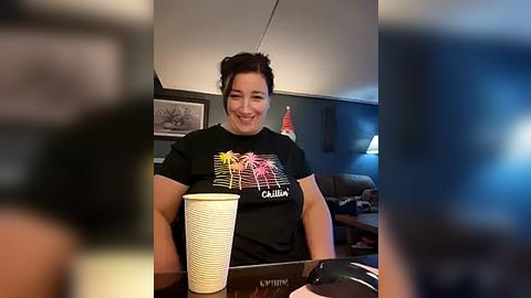 Media: A video of a smiling woman with dark hair in a bun, wearing a black T-shirt with a colorful graphic design, holding a white paper cup, seated at a desk in a dimly lit room with framed art and an American flag.