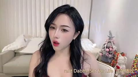 Media: Video of an East Asian woman with long black hair, fair skin, and red lipstick, wearing a sleeveless top, sitting in a modern living room with a decorated Christmas tree.