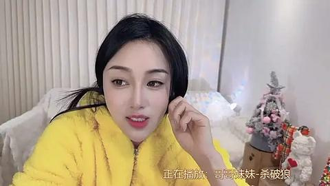 Media: Video of an East Asian woman with long black hair, wearing a yellow robe, smiling and holding her chin. Background features a Christmas tree, white walls, and vertical blinds.