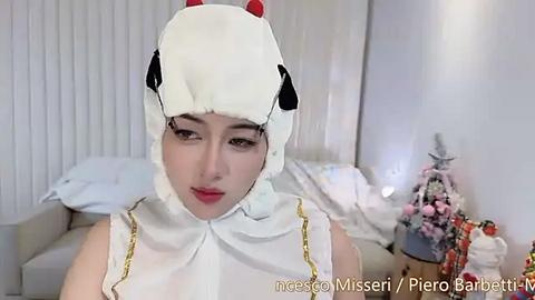 Media: Video of a young Asian woman in a white, fluffy cow costume, standing in a brightly lit bedroom with white vertical blinds. The room features a bed, a Christmas tree, and a small gift box.