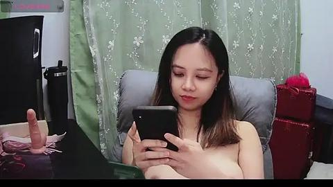 Media: Video of an East Asian woman with long, straight black hair, seated nude on a gray chair, texting on a smartphone, in a room with green floral curtains and red leather chair.