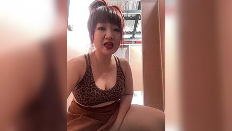 Media: Video of an Asian woman with light skin, brown hair in a high ponytail, wearing a leopard-print bra and brown skirt, sitting in a doorway with a blurred background.