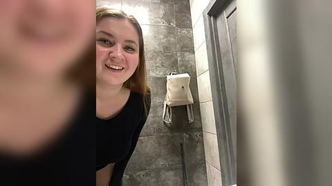 Media: Video of a smiling woman with light skin and straight, shoulder-length brown hair, wearing a black T-shirt, standing in a tiled bathroom with a beige hand towel dispenser on the wall.