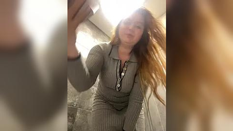 Media: Video of a plus-sized woman with long, wavy red hair, wearing a grey ribbed jumpsuit, smiling and taking a selfie in a dimly lit bathroom with a tiled floor.