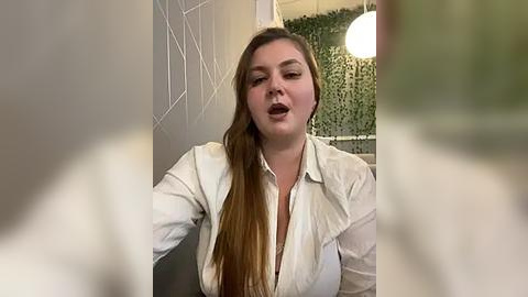 Media: A video of a fair-skinned woman with long, straight brown hair, wearing a white button-up shirt, laughing with her mouth open, in a modern indoor setting with white walls and a hanging lamp.