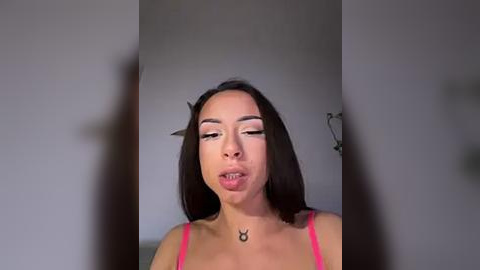 Media: Video of a young woman with medium brown skin and straight black hair, wearing a pink spaghetti strap top. She's licking her lips and has a small tattoo on her neck. Background is a plain, gray wall.