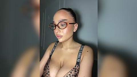 Media: Video of a fair-skinned woman with large breasts, wearing leopard print bra, dark-framed glasses, and wet hair, standing against a plain gray wall, slightly blurred background.