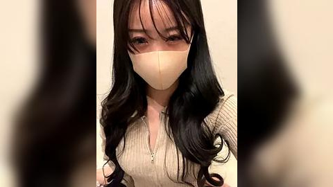 Media: Video of an East Asian woman with long, wavy black hair, wearing a beige face mask and a ribbed beige sweater. The background is blurred, focusing on her face.
