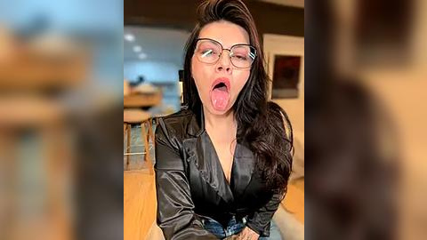 Media: Video of a woman with long black hair, wearing glasses, black satin jacket, and jeans, sticking her tongue out playfully. Indoor setting with blurred background, modern furniture.