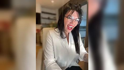 Media: Video of a smiling woman with long dark hair, wearing glasses and a white blouse, gesturing with her hands in a modern, well-lit room.