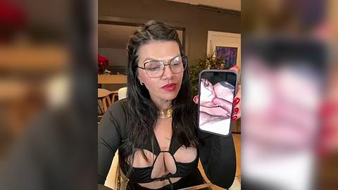 Media: Video of a woman with long dark hair, wearing glasses and a revealing black top with cutouts, taking a selfie in a cozy, dimly lit room.