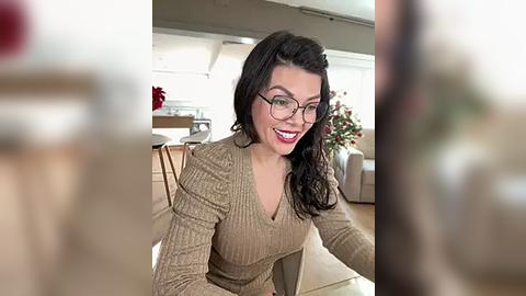 Media: Video of a smiling woman with long, dark hair, wearing glasses and a beige knit top, seated in a modern living room with a Christmas tree in the background.