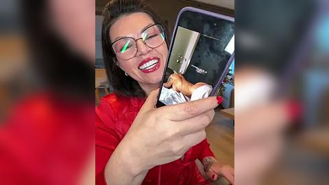 Media: Video of a woman with glasses, red lipstick, and a red top, holding a smartphone with a shiny case, winking and smiling. Background is blurry, suggesting she's taking a selfie.