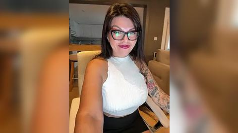 Media: Video of a tattooed woman with glasses and red lipstick, wearing a white ribbed tank top and black high-waisted shorts, seated in a modern living room.