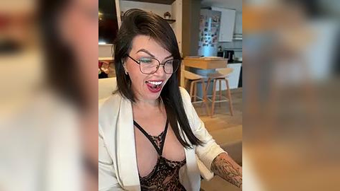 Media: Video of a middle-aged woman with fair skin, black hair, glasses, and a white blazer over a black lace bra, laughing in a modern kitchen with wooden stools and a fridge.