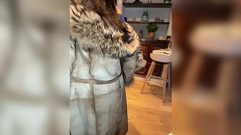 Media: Video of a person in a gray fur coat, standing in a modern kitchen with wooden floors, a white table, and shelves filled with glassware.