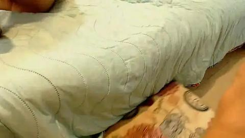 Media: Video of a person lying on a light-colored, quilted bedspread with visible wrinkles, showcasing a tan arm and leg. The bedspread covers a tan and brown patterned rug.