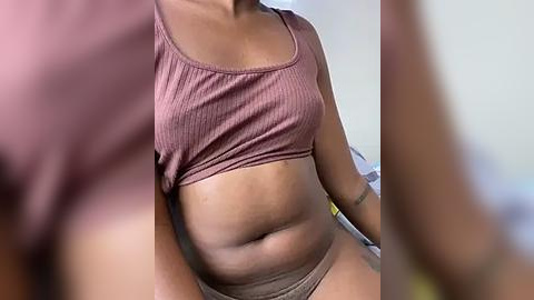 Media: A video of a plus-sized Black woman with medium-brown skin, wearing a ribbed mauve crop top and beige panties, sitting indoors with a blurred background.