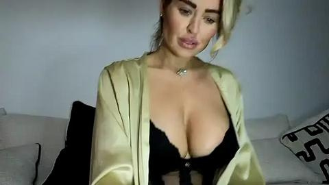 Media: Video of a curvy, light-skinned woman with large breasts, wearing a black lace bra and a silky, light green robe, seated on a gray couch, indoors, with a minimalist background.