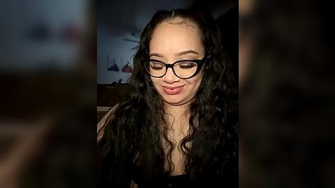 Media: Video of a smiling woman with curly black hair, wearing black-framed glasses, and a dark top, set against a dimly lit, blurred background.