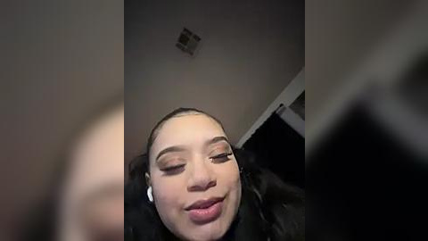 Media: Video of a young woman with light skin, dark hair, and brown eyes, smiling, wearing black headphones, in a dimly lit room with a small light fixture on the ceiling.