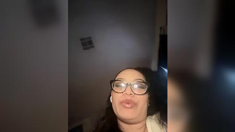 Media: Video of a woman with light skin, wearing glasses, making a kissy face in a dimly lit room.