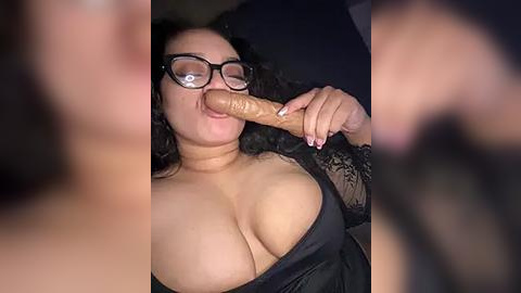 Media: A video of a curvy woman with glasses and long black hair, wearing a black lace top, performing oral sex on a large, erect penis. The background is blurred and dark.