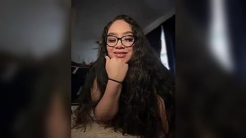 Media: Video of a young woman with long, curly black hair, wearing glasses, resting on her elbow in a dimly lit room with a window.