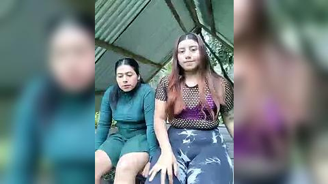 Media: Video of two women, one in a green bodysuit, the other in a black fishnet top and gray pants, sitting on a concrete floor with a green corrugated metal roof in the background.