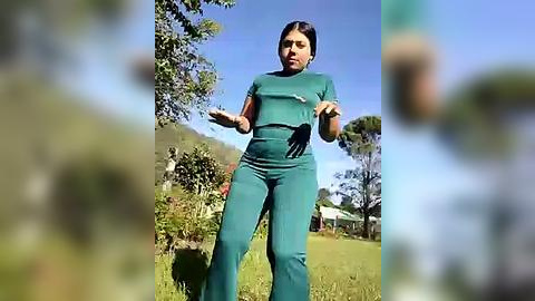 Media: Video of a woman in teal scrubs, black gloves, and a surgical mask, gesturing outdoors on a sunny day with blurred background of trees and grass.