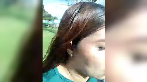 Media: Video of a woman with wet, dark brown hair partially covering her face, wearing a teal shirt, in a blurred, outdoor setting with green grass and trees.