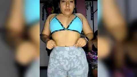 Media: A video of a plus-sized woman with medium brown skin, medium build, and dark hair, wearing a turquoise sports bra and light gray sweatpants, standing indoors with blurred background.