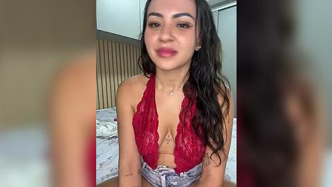 Media: A video of a Latina woman with long, wavy dark hair, wearing a red lace halter top and high-waisted jeans, smiling in a bedroom with wooden headboard and white bed.