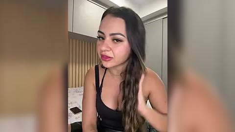 Media: Video of a young woman with long, dark hair, wearing a black tank top, smiling in a bedroom with a bed, wooden paneling, and closed closet doors.