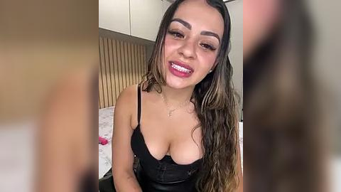 Media: Video of a smiling Latina woman with long brown hair, wearing a black lace bra and necklace, in a modern bedroom with wooden paneling.